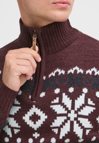Maglione a maglia bordeaux con colletto alto, caratterizzato da una zip e da motivi a fiocco di neve bianchi e teal sul petto e sulle spalle.