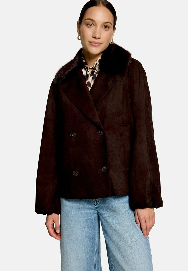 Femme portant une veste marron foncé à double boutonnage avec col en fourrure, une écharpe imprimé animalier, des boucles d'oreilles dorées en anneau et un jean bleu clair, debout sur fond blanc.
