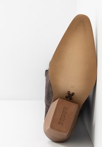 Bota de ante marrón de punta afilada apoyada contra una esquina blanca, mostrando suela texturizada, talla 37 y tacón bloque de marca.