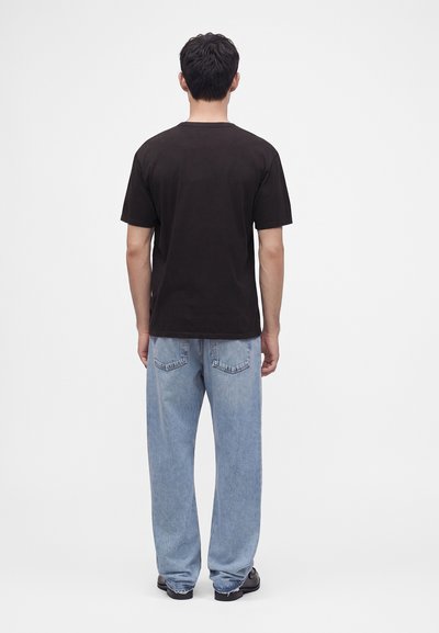 Homme debout, de dos, portant un t-shirt noir uni à manches courtes, un jean bleu clair et des chaussures noires sur fond blanc.