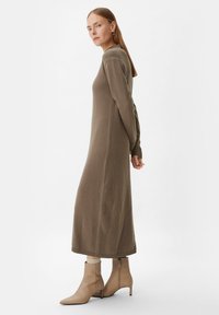 Langes, olivgrünes Strickkleid mit enganliegenden Ärmeln, seitlichen Schlitzöffnungen und geripptem Saum, kombiniert mit beigen Ankle-Boots mit spitzem Schuhzeichen.