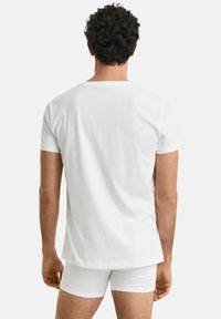 Man met krullend haar staat met zijn rug naar de camera, gekleed in een eenvoudig wit T-shirt met korte mouwen en witte aansluitende shorts, tegen een lichte achtergrond.