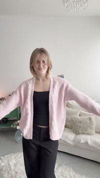 Mladá žena s blond vlasmi oblečená v ružovom kardigane a čiernom topu stojí s vystretými pažami v útulnej obývacej izbe.