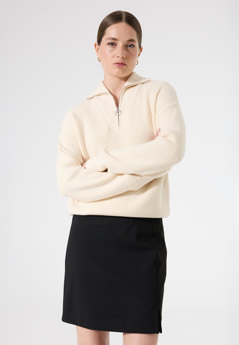 Suéter de punto acanalado color crema con media cremallera y tirador de anillo plateado, combinado con una falda negra ajustada. La textura parece suave y cálida.