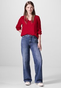 Rode blouse met lange mouwen en gathers op de schouders, gecombineerd met wijde blauwe denim jeans en lichtgekleurde canvas schoenen. Eenvoudige achtergrond.