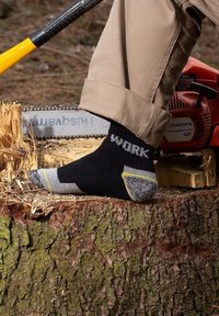 Calzini da lavoro neri con accenti grigi e gialli, punte rinforzate e la parola "WORK" stampata in bianco. Posati su un tronco vicino a una motosega.