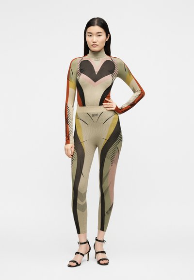 Γυναίκα στέκεται με το χέρι στη μέση, φοράει ένα εφαρμοστό ολόσωμο μαγιό με πολύχρωμο γεωμετρικό σχέδιο και μαύρα σανδάλια με ψηλά τακούνια σε απλό φόντο.