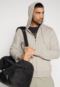 Ανοιχτό γκρι φούτερ με φερμουάρ και κουκούλα με κορδόνι, υλικό από βαμβακερό μείγμα, σε αντίθεση με μια μαύρη γυμναστηριακή τσάντα που διαθέτει λεπτομέρειες από δίχτυ.