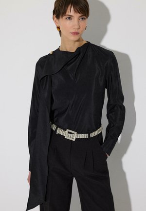 Ženska, oblečena v črno majico z dolgimi rokavi in asimetričnim ovratnikom, črne hlače in sijoč pas z okrasnimi kamni, stoji ob svetli podlagi.