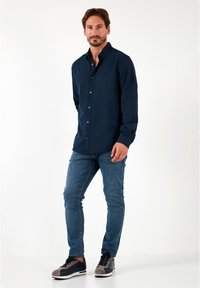 Camisa azul marino de botones, de mangas largas y con cuello, combinada con vaqueros ajustados azules y zapatillas oscuras con acentos grises. Diseño simple, sin estampados.
