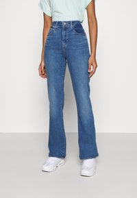 Femme portant un jean bleu taille haute, un T-shirt bleu clair rentré et des baskets blanches, se tenant devant un fond uni.