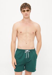 Zelené plavky s elastickým pásom, bielym šnúrkou a malým logom na nohavici. Model nosí slnečné okuliare a je bez trička.