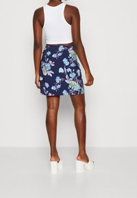 Žena v bílém bezrámovém topu, námořnickém modrém květinovém sukně a bílých platformových podpatcích, stojící na pozadí bez vzoru.