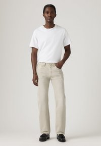Man standing facing forward wearing a white t-shirt, beige jeans, and black tassel loafers, with one hand in pocket against a plain background.