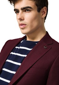 Burgundowa teksturowana marynarka nałożona na granatową koszulę w paski, z małym metalowym logo na klapie i dopasowanym kroju.