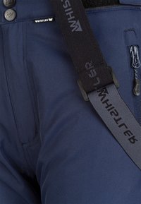 Gros plan sur un pantalon bleu avec une poche zippée, deux boutons et des bretelles noires de la marque Whistler attachées.