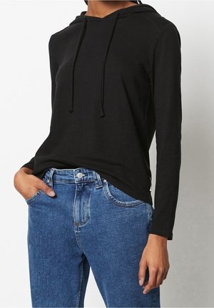 Persoon draagt een zwarte hoodie met lange mouwen en blauwe spijkerbroek, met één hand in de broekzak, tegen een effen achtergrond.