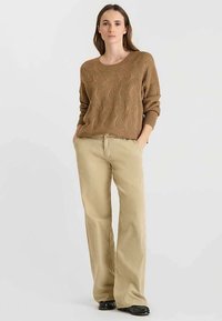 Maglione marrone a maglia con una trama pronunciata e scollo ampio, abbinato a pantaloni beige a gamba larga e calzature nere, posizionato contro uno sfondo chiaro.