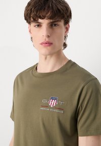 Camiseta de manga corta de color verde oliva, hecha de algodón, con un logo bordado que presenta un diseño de escudo y el texto: "GANT 1949 AMERICAN SPORTSWEAR."