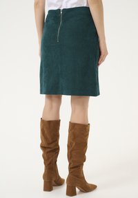 Jupe crayon en velours côtelé teal avec une fermeture éclair au dos ; longueur genou. Associée à des bottes en suède beige hauteur genou avec des talons épais.