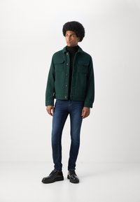 Chaqueta de tejido grueso verde con dos bolsillos frontales, botones marrones, combinada con jeans ajustados de color azul oscuro y zapatos negros con cordones.