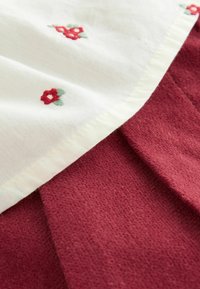 Bawełniana tkanina z białą górną częścią zdobioną czerwonym haftem kwiatowym; bordowa teksturowana dolna część z widocznym przeszyciem i fałdami.