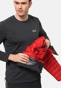 Rote und schwarze isolierte Jacke mit glänzender Textur, die ein pufferdesign aufweist und ein Logo auf der Brust hat. Das Modell hält die Jacke, während es ein dunkelblaues Langarmshirt trägt.