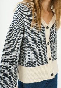 Cardigan a righe blu navy e crema con scollo a V, bottoni in legno e orlo a costine. Il motivo presenta forme geometriche su tutto il tessuto.