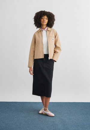 Donna con capelli ricci che indossa una giacca beige, camicia bianca, gonna midi nera e ballerine rosa pallido, in piedi su un tappeto blu.