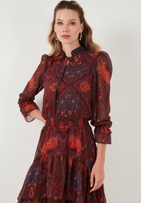 Punainen ja sininen paisley-kuvioinen mekko, jossa on pitkät hihat, joustava vyötärö ja röyhelöhelma. Valmistettu läpikuultavasta, keveästä kankaasta.