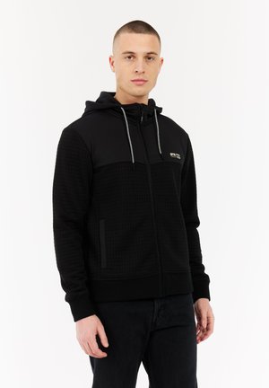 Jonge man met kort haar, gekleed in een zwart, gestructureerd zip-up hoodie met capuchon en trekkoorden, staande tegen een plain witte achtergrond.