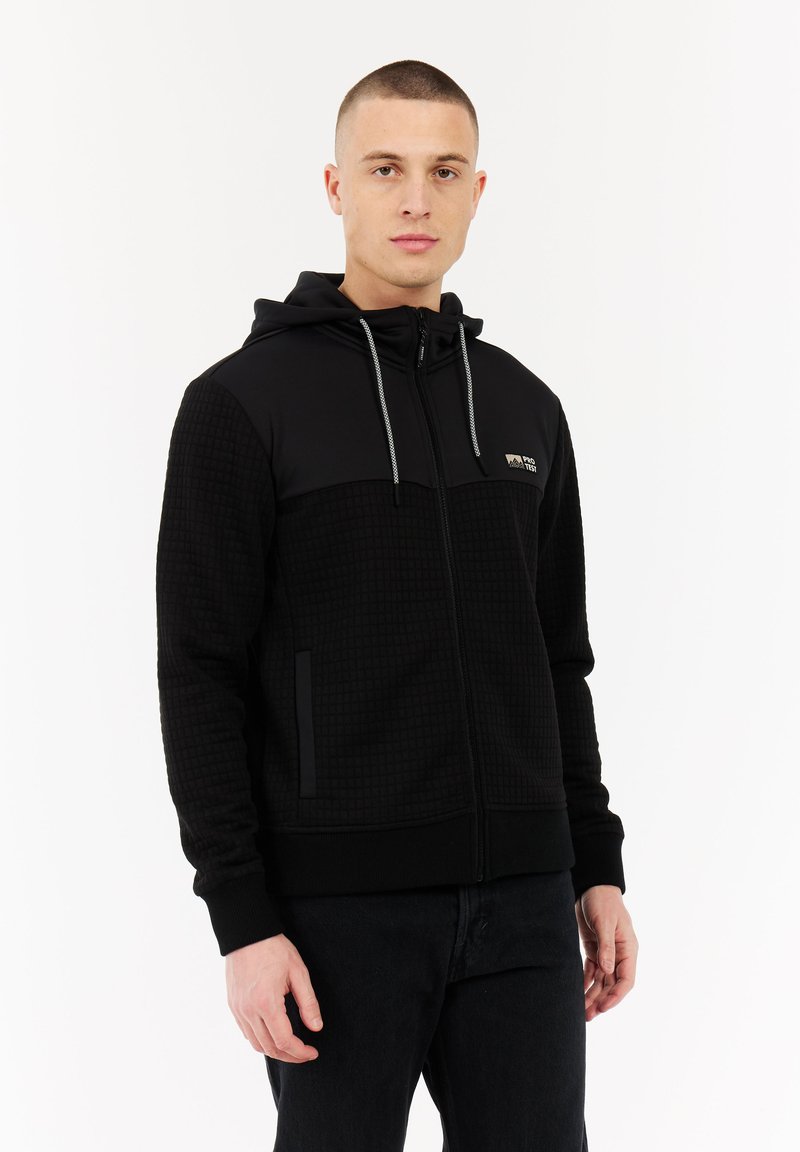 Jonge man met kort haar, gekleed in een zwart, gestructureerd zip-up hoodie met capuchon en trekkoorden, staande tegen een plain witte achtergrond.