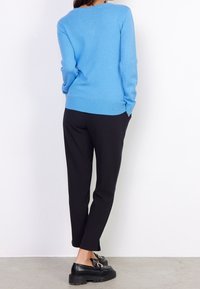 Frau in einem blauen Pullover, schwarzen engen Hosen und schwarzen Slippern, die mit den Händen in den Taschen vor einer weißen Wand steht.