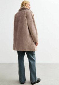 Cappotto in pelliccia sintetica di colore marrone chiaro, forma oversize con colletto alto. Indossato su jeans blu larghi, che mostrano un tessuto morbido e testurizzato.