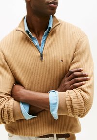Uomo che indossa un maglione beige a costine con cerniera corta sopra una camicia azzurro chiaro con bottoni, braccia incrociate, inquadratura dal naso fino alla vita.