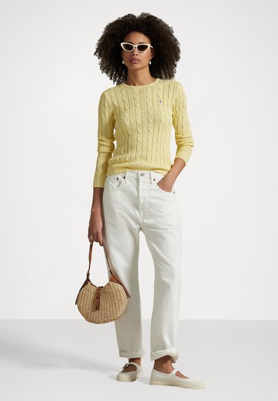 Sweater amarelo de malha com padrões de cordas, combinado com calças de ganga brancas de cintura alta, uma bolsa redonda de vime e sapatos slip-on de cores claras.
