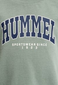 Grön sweatshirt med fet marinblå text "HUMMEL" och vit text "SPORTSWEAR SINCE 1923" under, med ribbad krage och struktur.