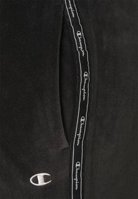 Svarta fleecebyxor med sidofickor, med vitt Champion-logotypdetalj och mönstrad tejp längs sömmen.