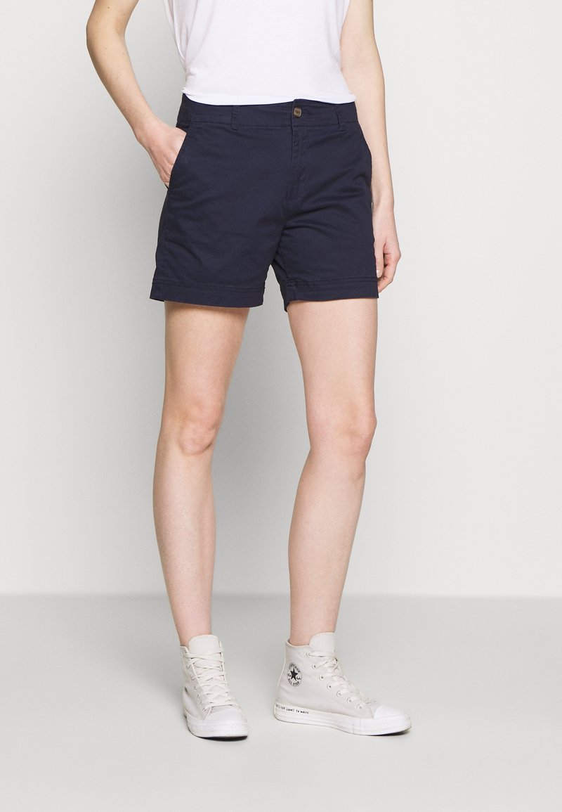 Personne portant un short bleu marine, un t-shirt blanc et des baskets montantes blanches, debout avec une main dans la poche sur un fond uni.