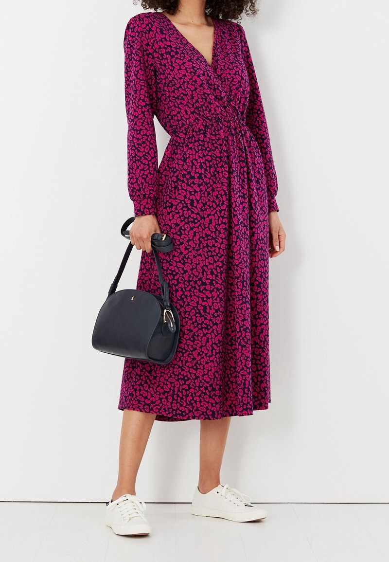 Femme portant une robe midi à motifs violets et roses à manches longues, des baskets blanches, et tenant un sac à main noir à côté d'elle.
