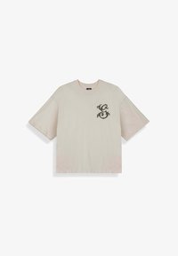 Camiseta beige de manga corta extragrande con cuello redondo, con un pequeño diseño ornamental oscuro en forma de "S" en el pecho izquierdo.