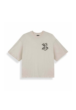 Camiseta beige de manga corta extragrande con cuello redondo, con un pequeño diseño ornamental oscuro en forma de "S" en el pecho izquierdo.