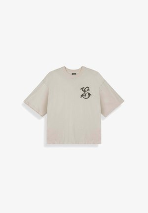 Beżowa, luźna koszulka z krótkim rękawem i okrągłym dekoltem, z małym ciemnym ozdobnym wzorem "S" na lewej piersi.