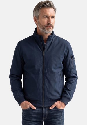 Mann mit grauen Haaren und Bart, der eine marineblaue Jacke mit Reißverschluss und dunkle Jeans trägt, steht mit den Händen in den Taschen und schaut nach rechts.