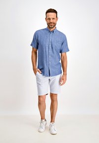 Camicia azzurra a maniche corte con bottoni e taschino sul petto, abbinata a pantaloni corti a righe e sneakers bianche su uno sfondo neutro.
