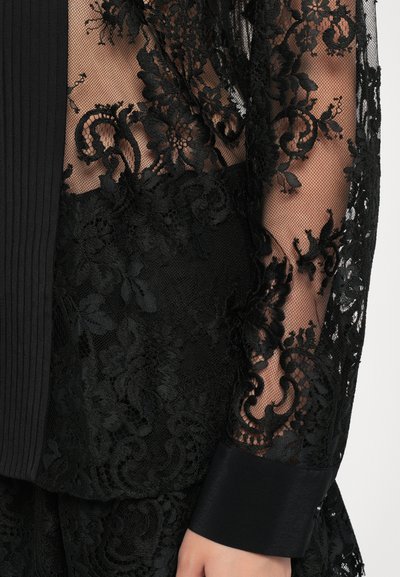 Svart bluse i blonder med intrikate blomstermønstre, et teksturert mesh-overlag og satengmansjetter med glatt finish.