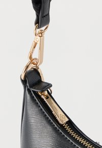 Γωνία τσάντας από μαύρο δερμάτινο με υφή, με χρυστό φερμουάρ και κούμπωμα προσαρτημένα σε μαύρο λουράκι σε λευκό φόντο.