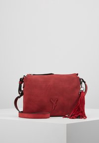 Sac à bandoulière en cuir perforé rouge avec une sangle détachable, fermeture à rabat et un porte-clés à pompon rouge sur un piédestal blanc contre un fond gris.