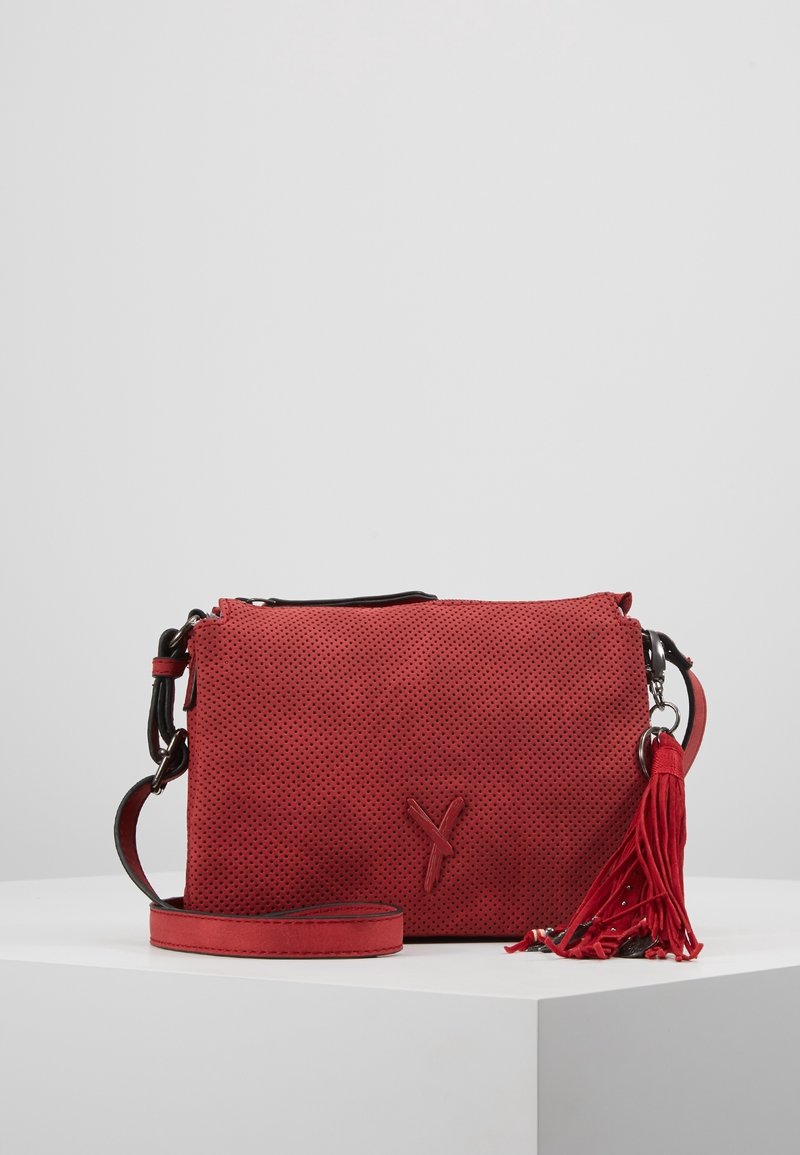 Sac à bandoulière en cuir perforé rouge avec une sangle détachable, fermeture à rabat et un porte-clés à pompon rouge sur un piédestal blanc contre un fond gris.