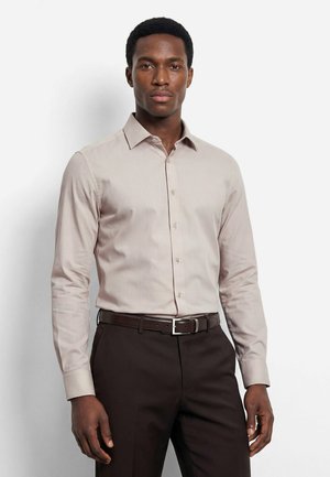 Man draagt een lichtbeige overhemd met knopen, in donkerbruine broek met bijpassende riem, staand tegen een effen achtergrond.
